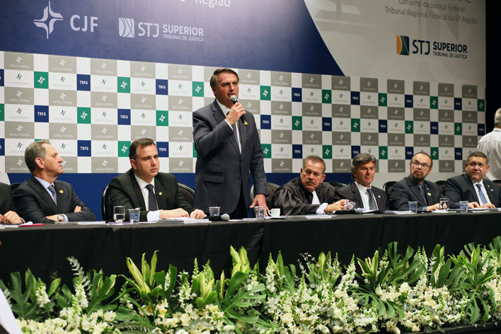 Foto horizontal de mesa de honra mostrando 7 pessoas uma delas em pé falando no microfone.