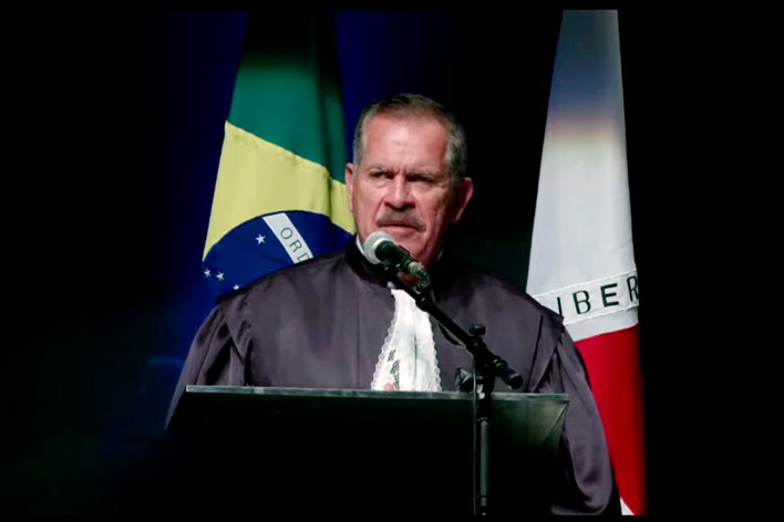 Foto de perfil de um homem branco cabelo curto e bigode vestindo toga. Falando ao microfone em um púlpito com bandeira do brasil e de minas gerais ao fundo.