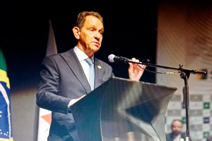 Foto de perfil de um homem branco cabelo curto, vestindo terno azul escuro, com gravata e blusa azul clara. Falando ao microfone em um púlpito com bandeira do brasil e de minas gerais ao fundo.